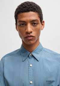Lichtblauw button-up overhemd met een puntige kraag, één zak met geborduurd logo, soepele textuur en subtiele glans.