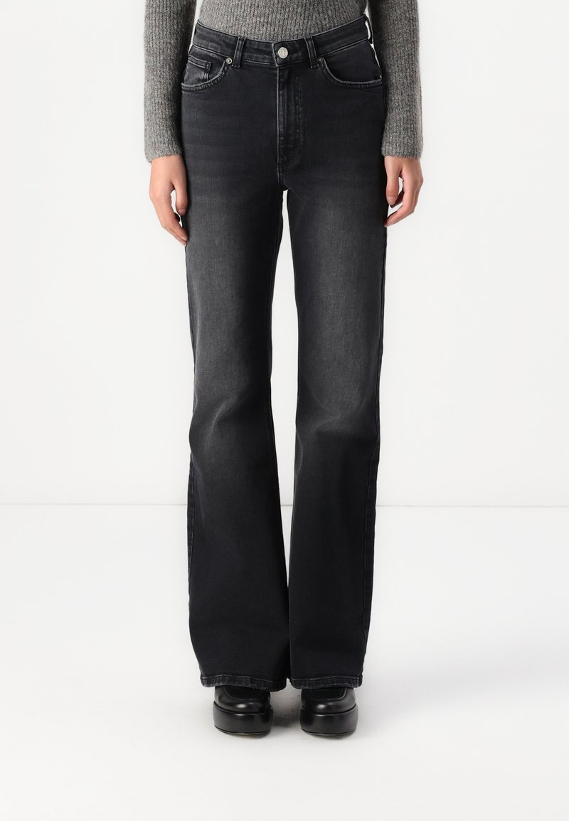 Personne portant un jean noir taille haute à jambes larges avec un bouton et une fermeture éclair, associé à des chaussures noires et un haut gris à manches longues.
