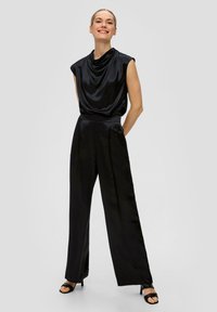 Haut sans manches en satin noir avec un décolleté drapé, associé à un pantalon noir taille haute et large. Accentué par des sandales à talons noires.