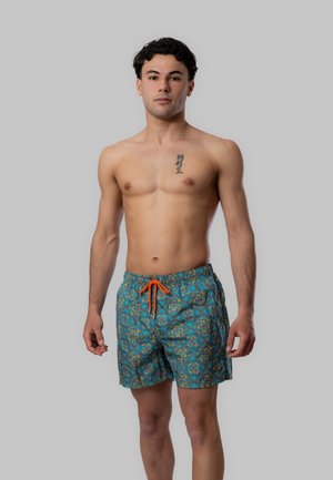 Giovane uomo in piedi a torso nudo che indossa pantaloncini da bagno blu con motivi e coulisse arancione, con tatuaggio sul petto con caratteri coreani.