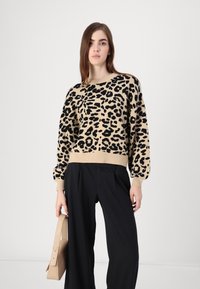 Pulover s uzorkom leoparda u bež i crnoj boji, s okruglim izrezom i dugim rukavima. Uparen s crnim hlačama širokih nogavica i bež torbicom.