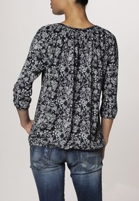 Blusa floral azul marino con patrones blancos, escote fruncido, mangas largas y corte relajado. Combinada con vaqueros azules con costuras decorativas.