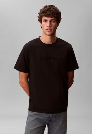 Jaunas vaikinas su garbanotais plaukais, dėvintis juodą Calvin Klein marškinėlį ir pilkus džinsus, stovintis su rankomis už nugaros priešais vientisą foną.