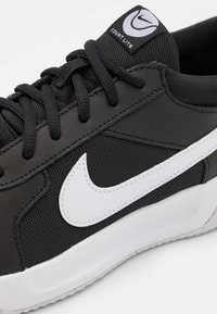 Svart sportskor med ovandel i mesh, vit Nike-swoosh och en texturerad sula. Rund tå och framträdande snörningssystem.