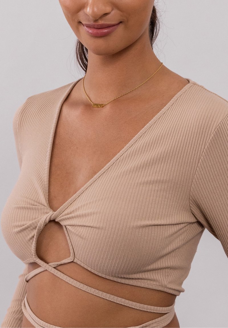 Femme portant un crop top beige côtelé à manches longues avec un nœud central et une découpe, associé à un délicat collier en or.