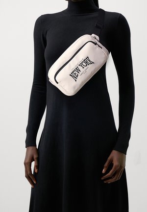 Persoon in een zwarte jurk met lange mouwen die een beige crossbodytas draagt met de tekst "New York College Team Varsity League" in het zwart.