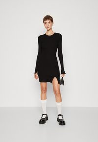Robe noire à manches longues côtelées avec une fente sur le côté, présentant une coupe ajustée et une texture douce. Associée à des chaussettes blanches montantes et des chaussures noires épaisses.