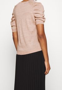 Lichtroze gebreide trui met drie-kwart trompetmouwen, aangesloten zoom en een gladde textuur, gedragen over een zwarte geplooide rok.