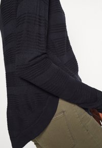 Pull en maille texturée bleu marine avec rayures horizontales, présentant un col rond et des poignets côtelés, associé à un pantalon vert olive.