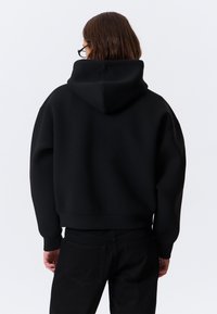 Hanorac scurt negru cu glugă, realizat dintr-un material moale. Prezintă umeri căzuți și manșete ribbed. Poartă pantaloni negri.