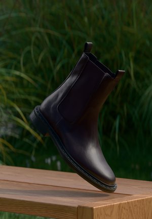 Stivale Chelsea in pelle marrone scuro con pannelli laterali elastici e linguette, posizionato su una superficie in legno con sfondo di vegetazione sfocata.