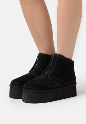 Black suede platform boots with a rounded toe and twin zip closures on the front, featuring a thick, textured base for added height.