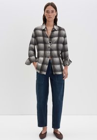 Camicia a scacchi bianca e nera con colletto e bottone circolare, abbinata a pantaloni in denim blu rilassati e scarpe slip-on marroni.