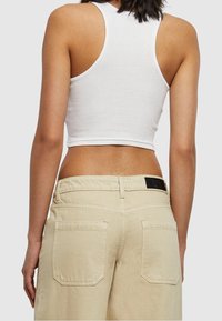 Débardeur blanc court avec des fines bretelles et un pantalon large beige clair avec deux poches arrières et une étiquette noire sur la taille.