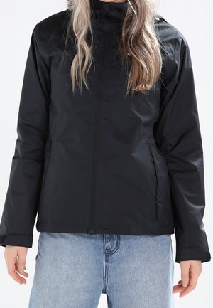 Femme portant une veste noire zippée avec des poches latérales zippées et un jean en denim bleu clair, se tenant devant un fond uni.