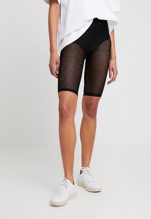 Personne portant un short de vélo noir transparent à motifs, une chemise blanche oversize et des baskets blanches, debout sur un fond clair uni.