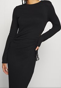 Musta, pitkähihainen mekko ribbikankaasta, jossa on sivurypytys ja säädettävät narut helmassa. Sujuva pinta, tiukka malli.