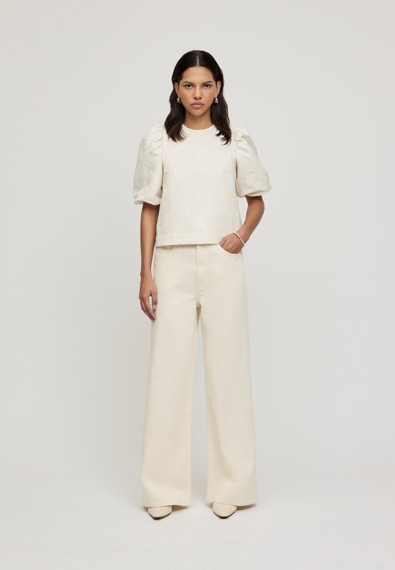 Top strutturato color crema con maniche a sbuffo, abbinato a pantaloni a gamba larga color crema e scarpe beige, caratterizzato da motivi sottili e linee pulite.