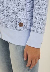 Svijetloplavi dukserica sa zipom koja ima uzorak dijamanta i točkica, rebraste manžetne i tamniju oznaku na rubu. Nosena preko svijetlog denima.
