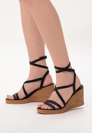 Ben i sorte sandaletter med kilehæl i vævet brunt på hvid baggrund.