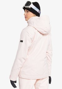 Lichtroze waterdichte skijas met een kap, zwarte accenten op de schouder en een soepele textuur. Draag met zwarte handschoenen voor extra warmte.