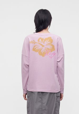 Personne portant une chemise lavande à manches longues avec une grande fleur d’hibiscus orange et un texte rose "Roxy" dans le dos, assortie à un pantalon cargo gris.