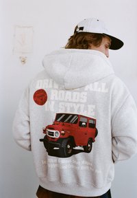 Šedá mikina s kapucí s červenou grafikou vintage Toyota Land Cruiser, textem "JEĎTE VŠEMI CESTAMI VE STYLU" a kulatou nášivkou s logem Toyota.