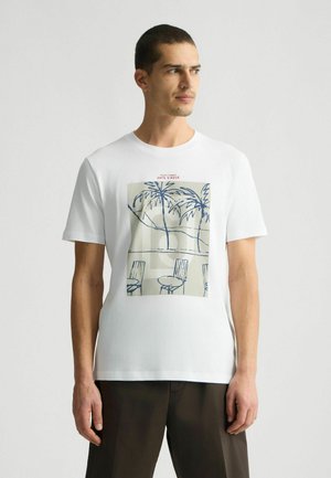 Camiseta de algodón blanca con un estampado gráfico de palmeras y sillas en azul y beige. Cuello redondo y mangas cortas.