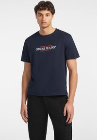 Tamsiai mėlynas medvilninis marškinėliai, kuriuose yra centrinis raudonas ir baltas logotipas "GUESS JEANS" su užrašu "AMERICAN TRADITION" viršuje ir "ORIGINAL DESIGNS" apačioje.