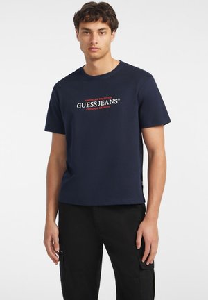 Námořnické modré bavlněné tričko s centrálním červeným a bílým logem s textem "GUESS JEANS" s nápisem "AMERICAN TRADITION" nad ním a "ORIGINAL DESIGNS" pod ním.
