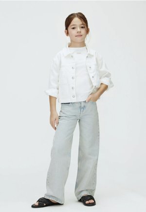 Giovane ragazza con capelli castani, giacca di jeans bianca, camicia bianca, jeans larghi azzurri chiari e sandali neri, in piedi con una mano in tasca.