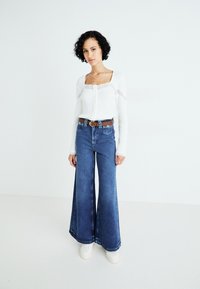 Witte ribgebreide lange mouw top met kantdetails, gecombineerd met wijde blauwe denim jeans en witte platform schoenen, met een bruine riem.