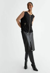 Gilet nera con zip e maniche beige, con tasca frontale e dettaglio della fibbia, abbinato a una gonna in pelle nera e stivali a coscia in pelle testurizzata.
