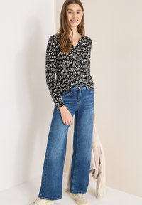 Sort langærmet top med beige abstrakt mønster, kombineret med vidde-ben blå denimjeans og cremefarvede sneakers, båret med en lys jakke.