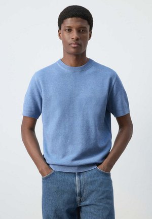 Mladý muž stojí s rukami v vreckách, nosí svetlomodrý krátky sveter a modré džínsy, neutrálne pozadie.