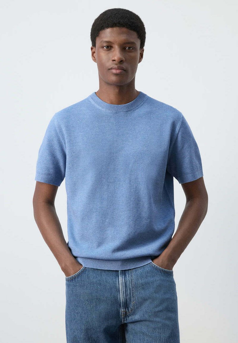 Jeune homme debout, les mains dans les poches, portant un pull léger bleu clair à manches courtes et un jean bleu, fond neutre.