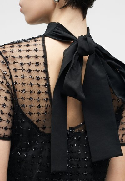 Black dress featuring sheer fabric with floral beadwork, a deep back neckline, and a large ribbon tie at the collar.