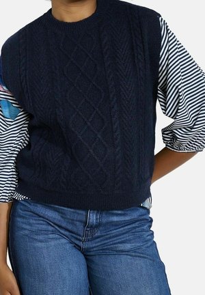 Personne portant un gilet en tricot bleu marine par-dessus un chemisier rayé à manches longues bouffantes et un jean bleu, debout avec une main sur la hanche.
