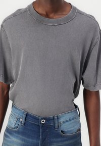 Osoba v vybledlé šedé polokošili s krátkým rukávem, zasunuté do vybledlých modrých džínů, na kterých jsou viditelné knoflíky a páskové smyčky.