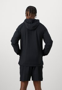 Musta kevyt takki hupulla, pitkät hihat ja suora leikkaus, yhdistettynä mustiin lyhyisiin housuihin. Sileä pinta, minimalistinen muotoilu.