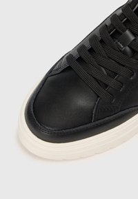 Zapatilla negra de cuero con parte superior texturizada, cordones negros planos y suela de goma blanca. Presenta un sutil detalle de costura a lo largo del borde.