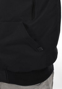 Veste noire avec une poche zippée et un tissu texturé. Comprend une taille côtelée et un petit logo près de la poche.