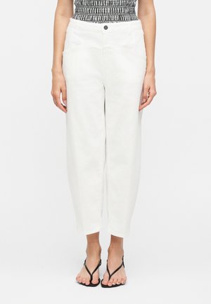 Kvinde iført hvide korskårne bukser med høj talje, sort-hvid rynket top og sorte sandaler med stropper, stående mod en ensfarvet hvid baggrund.