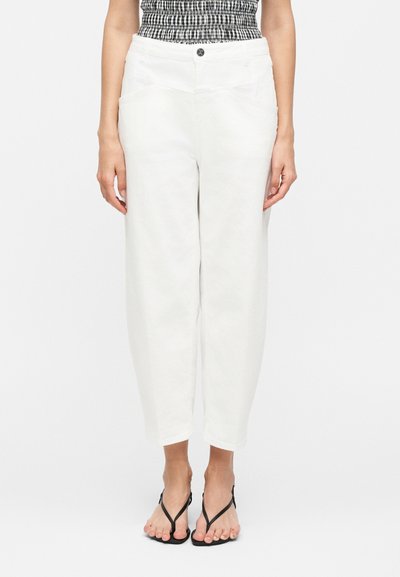 Femme portant un pantalon blanc court taille haute, un haut smocké noir et blanc, et des sandales noires à brides, debout devant un fond blanc uni.