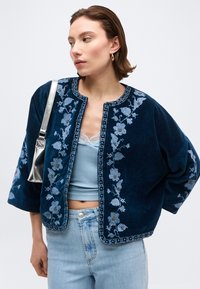 Γυναίκα που φορά μπλε κεντημένο cropped σακάκι, ανοιχτό γαλάζιο τοπ από δαντέλα, τζιν ανοιχτού χρώματος, χρυσούς κρίκους σκουλαρίκια και ασημένια τσάντα ώμου.