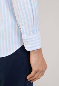 Camicia azzurro chiaro con righe verticali rosa e bianche. Presenta un polsino con bottone e un orlo arrotondato. Texture liscia.