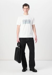 Witte katoenen t-shirt met golfpatroon in blauw en roze, gecombineerd met zwarte rechttoe-rechtaan jeans en een zwarte tote bag.