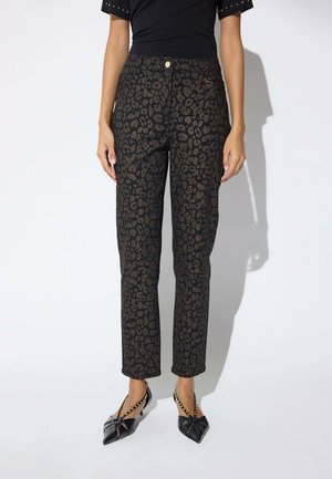 Persona que lleva pantalones rectos con estampado de leopardo negro y marrón combinados con zapatos negros puntiagudos con tachuelas doradas y correas.