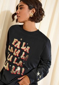 Melna sporta krekla ar sarkaniem un zeltainiem spīdīgiem burtiem un zvaigznēm. Materiāls ir gluds, ar apaļu kakla izgriezumu un ribotām piedurknēm.