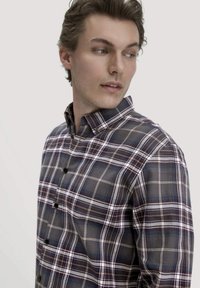 Jonge man die zijwaarts kijkt, gekleed in een knooploze, meerkleurige plaid shirt met een kraag, tegen een effen lichte achtergrond.